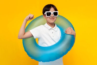 Güneş gözlüklü on iki yaşındaki çocuk şişme bir yüzme yüzüğü tutuyor ve sarı izole edilmiş arka planda güç ve başarı göstergesi gösteriyor. Plajda yazın doğan çocuk pazılarını gösteriyor.
