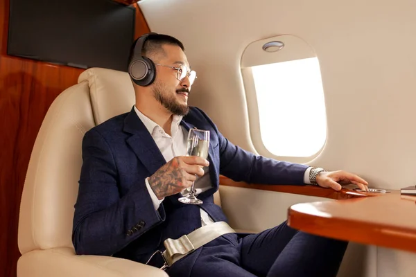 stock image successful Asian businessman flies in private luxury jet with glass of champagne and listens to music in headphones, Korean entrepreneur in suit rests and relaxes in airplane, luxury lifestyle