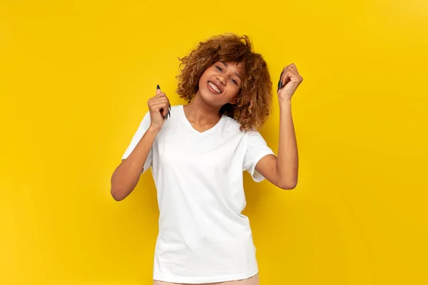 stock image young curly american girl dancing to the music on yellow isolated background, african woman in white t-shirt with her eyes closed moves with her hands up