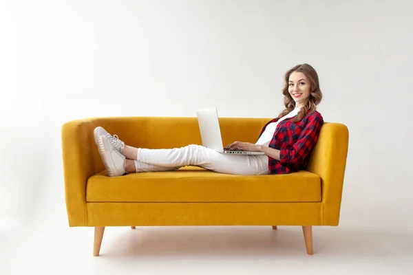 若いかわいい女の子は柔らかい快適なソファの上にあり ラップトップを使用しています 女性タイプオンライン上のコンピュータ上の黄色のソファの上に白い隔離された背景 — ストック写真