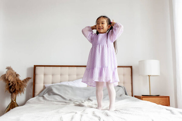 little Asian girl in dress rejoices and jumps on the bed at home, Korean child in festive outfit dances in the room on the bed