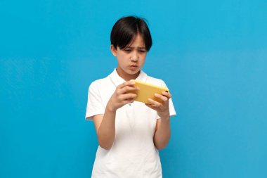 12 yıllık Asyalı çocuk mavi izole edilmiş arka planda akıllı telefondan video oyunu oynuyor, Koreli çocuk oyuncu mobil online oyun odaklı