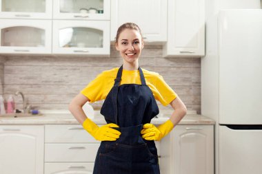 Önlüklü, eldivenli, neşeli ev hanımı evde temizlik yapıyor ve gülümsüyor. Üniformalı kadın, mutfağın arka planında dikiliyor.