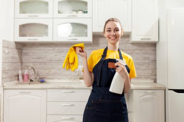Önlüklü, eldivenli, neşeli ev hanımı evde temizlik yapıyor ve gülümsüyor. Üniformalı kadın deterjan tutuyor ve mutfağı temizliyor.