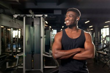 Genç Afrikalı Amerikalı adam koyu renk spor salonunda kollarını kavuşturup gülümsüyor fitness antrenörü fitness kulübünde duruyor ve reklamlar mekanı kopyalıyor.