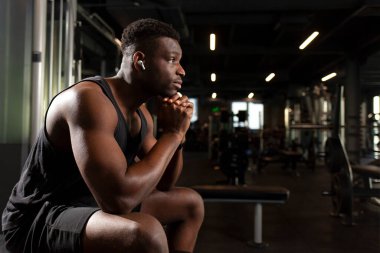 young athletic african american man sits in dark gym and thinks, pensive athletic guy rests and looks forward in fitness club, portrait of athlete in training clipart