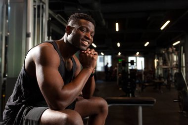 Genç atletik Afrikalı Amerikalı adam koyu renk spor salonunda oturuyor ve gülümsüyor, atletik adam dinleniyor ve fitness kulübündeki kameraya bakıyor, antrenmandaki sporcunun portresi.
