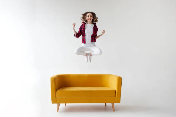 stock image young cute girl jumps and rejoices in victory on soft comfortable sofa, woman celebrates success and good luck on yellow couch on white isolated background