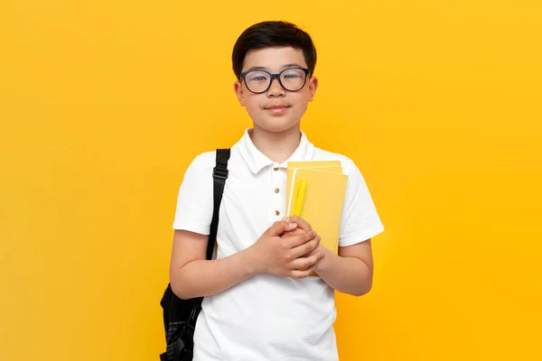 10岁的亚裔男生戴着眼镜 背着背包 手里拿着黄色背景的书 回到学校 满脸笑容 — 图库照片
