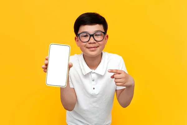 stock image asian little boy of ten years old in glasses shows blank screen of smartphone on yellow isolated background and smiles, korean child advertises copy space on the display of mobile phone