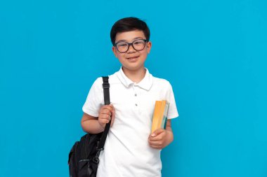 Sırt çantalı ve mavi arka planda gülümseyen küçük Asyalı okul çocuğu, Koreli çocuk, tecrit edilmiş arka planda çalışmak için okula gidiyor.