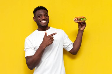 Beyaz tişörtlü Afro-Amerikan adam elinde büyük bir burgerle çizburgerciye işaret ederek sarı izole edilmiş arka planda fast food öneriyor ve reklamını yapıyor.
