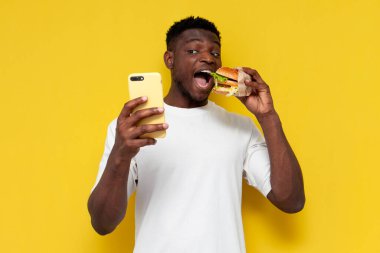 Beyaz tişörtlü mutlu Afro-Amerikan adam elinde büyük bir hamburger tutuyor ve akıllı telefon kullanıyor. Fast food yiyor ve sarı izole edilmiş arka planda internet üzerinden yemek siparişi veriyor.