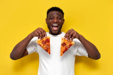 Beyaz tişörtlü mutlu Afro-Amerikan adam iki dilim pizza tutuyor ve sarı izole arka planda gülümsüyor, genç komik adam fast food yiyor ve bağırıyor.