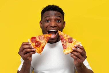 Beyaz tişörtlü mutlu Afro-Amerikan adam iki dilim pizza tutuyor ve sarı izole arka planda gülümsüyor, genç komik adam fast food yiyor ve bağırıyor.