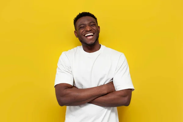 stock image happy african american man in white t-shirt stands with his arms crossed on yellow isolated background, toothy guy laughs and looks at the camera