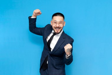 Başarılı iş adamı takım elbiseli ve gözlüklü Asyalı zaferi kutluyor ve mavi yalıtılmış arka planda kutluyor. İş kıyafetleri içindeki Koreli girişimci iyi şansa seviniyor.