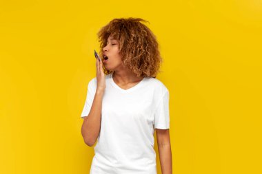 Genç yorgun kıvırcık Amerikalı kız elini sarı izole arka planda uzatır ve ağzını kapatır. Beyaz tişörtlü Afrikalı uykulu kadın uykusuzluktan muzdarip olur.