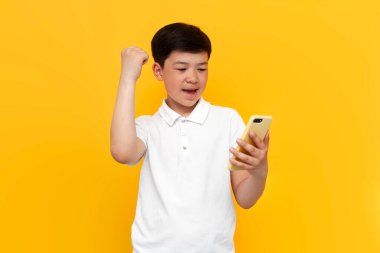 On yaşındaki beyaz tişörtlü Asyalı çocuk, akıllı telefon kullanıyor ve sarı izole edilmiş arka planda zaferle seviniyor. Cep telefonlu Koreli çocuk kazananı, iyi şans ve başarıyı kutluyor.