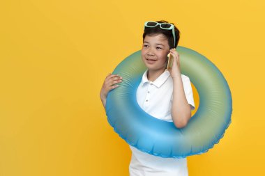On yaşındaki Asyalı küçük bir çocuk, şişme yüzme yüzüğüyle sarı izole edilmiş arka planda telefonla konuşuyor, yaz tatilinde Koreli çocuk cep telefonuyla iletişim kuruyor.