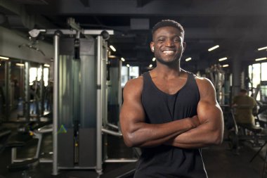 Genç Afrikalı Amerikalı adam koyu renk spor salonunda kollarını kavuşturup gülümsüyor fitness antrenörü fitness kulübünde duruyor ve reklamlar mekanı kopyalıyor.
