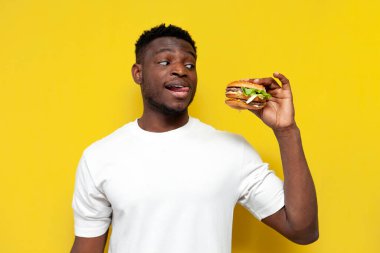 Beyaz tişörtlü Afro-Amerikan adam büyük bir burger tutuyor ve sarı izole arka planda dudaklarını yalıyor, genç adam fast food gösteriyor ve