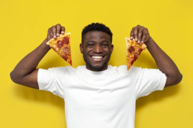 Beyaz tişörtlü neşeli Afro-Amerikan adam iki dilim pizza tutuyor ve sarı izole edilmiş arka planda gülümsüyor, fast food 'lu adam dişlek gülümseme gösteriyor.