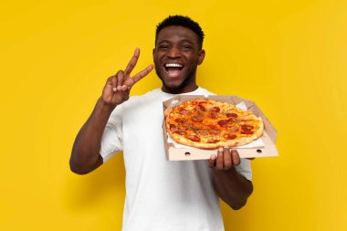 Beyaz tişörtlü neşeli Afro-Amerikan adam pizza kutusunu tutuyor ve sarı izole edilmiş arka planda barış jesti gösteriyor, adam fast food reklamları yapıyor ve hoş geldin diyor.