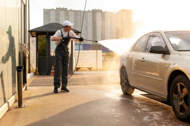 Üniformalı genç adam araba yıkama işçisi arabayı hortumla yıkıyor, tulumlu adam dışarıda araba temizleme servisinde çalışıyor.