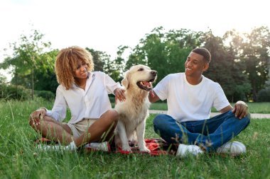 Genç Afro-Amerikan çifti ve köpeği yeşil çimenlerde oturuyor, kıvırcık kadın ve genç adam Golden Retriever ile birlikte, doğada evcil hayvanı olan mutlu bir aile.