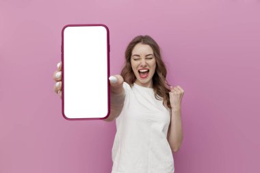 Beyaz tişörtlü genç tatlı kadın boş akıllı telefon ekranı gösteriyor ve pembe izole edilmiş arka planda zaferle seviniyor. Kazanan kız telefondaki fotokopi alanının reklamını yapıyor.