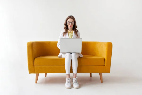 若いビジネスマンスーツの女性は快適な柔らかいソファに座って ラップトップを使用して 正式な摩耗の女の子は白い隔離された背景に黄色のソファの上にコンピュータに入力されています — ストック写真