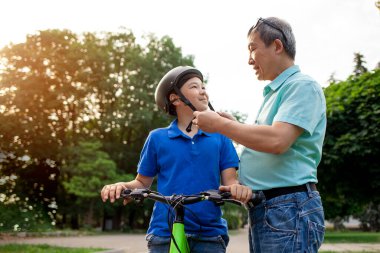 Baba, Asyalı oğlunun kafasına koruyucu başlık takıyor Koreli çocuk yazın babasıyla birlikte parkta bisiklet sürüyor, baba çocuğa bakıyor.