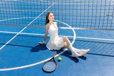 Beyaz üniformalı genç bayan tenisçi dışarıda mavi kortta oturuyor ve dinleniyor.