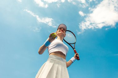 Beyaz üniformalı kız tenisçi tenis kortunda raket ve top tutuyor, kadın sporcu açık havada gökyüzüne karşı tenis oynuyor