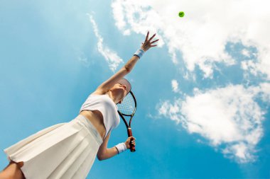 Beyaz üniformalı kız tenis oyuncusu raket tutuyor ve tenis kortunda topu fırlatıyor, bayan sporcu dışarıda gökyüzüne karşı tenis oynuyor.