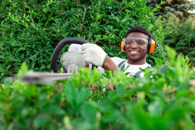 Üniformalı bahçe işçisi çalıları keser, gözlüklü ve kulaklıklı Afro-Amerikan adam bahçede elektrikli aletlerle, budama ağaçlarıyla ve çalılarla çalışır.