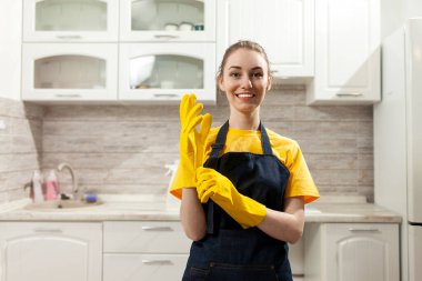 Önlüklü, eldivenli, neşeli ev hanımı evde temizlik yapıyor ve gülümsüyor. Üniformalı kadın, mutfağın arka planında dikiliyor.