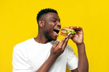 Beyaz tişörtlü Afro-Amerikan adam büyük burgeri ısırır ve sarı izole edilmiş arka planda sevinir, genç adam fast food yer ve sevinir, yakın plan