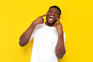 Neşeli Afrikalı Amerikalı adam ellerini sarı yalıtılmış arka planda dişlek beyaz gülüşüne doğrultur. Mutlu adam neşeli yüzünü gösterir.