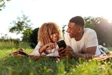 Afrika kökenli Amerikalı genç bir çift yazın parkta uzanıyor ve akıllı telefon kullanıyor. Erkek ve kadın cep telefonuna bakıyor ve doğada internetten yazı yazıyor.
