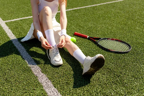 白いスポーツの若い女の子のテニス選手は彼女の靴を結ぶ上のスニーカーで緑のコート 女性コーチとテニスラケット — ストック写真
