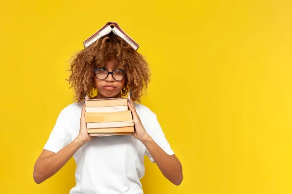 young tired curly american girl student in glasses holds books on yellow isolated background and is bored, african unhappy woman in white t-shirt is tired of studying and sad