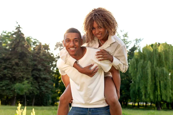 アフリカ系アメリカ人の幸せなカップルが夏に公園を歩いて笑顔で 男は彼の背中に女性を運び 自然の中で抱擁 自由と関係の概念 — ストック写真