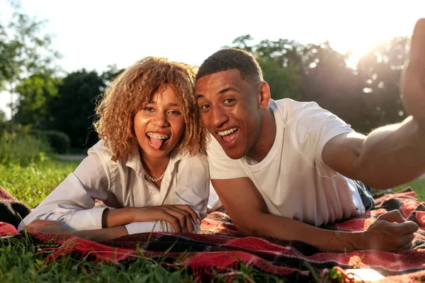 アフリカ系アメリカ人の若いカップルが夏の公園で草の上に横たわっており 自分自身を取り 男性と女性は自然の中でリラックスし 電話で自分自身の写真を撮る — ストック写真