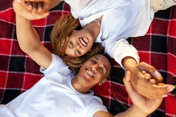 アフリカ系アメリカ人の若いカップルが毛布の上に横になって顔を上げて笑っている女性と男は手を取り合っている — ストック写真