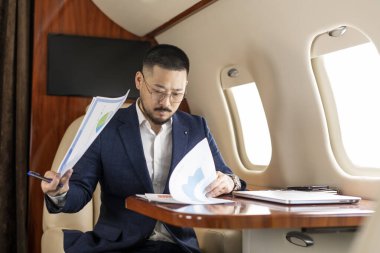 Takım elbiseli ve gözlüklü başarılı Asyalı işadamı özel jette oturuyor ve belgelerle çalışıyor. Koreli finansör, uçakta evrakları ve çizelgeleri kontrol ediyor.
