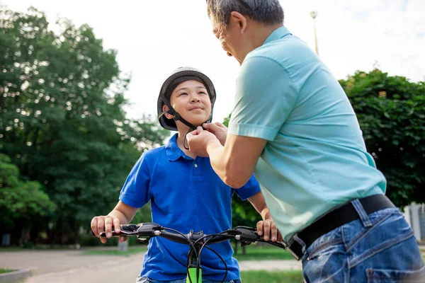 stock image father puts protective helmet on his Asian son's head, Korean boy rides bike with dad in the park in summer, father takes care of the child