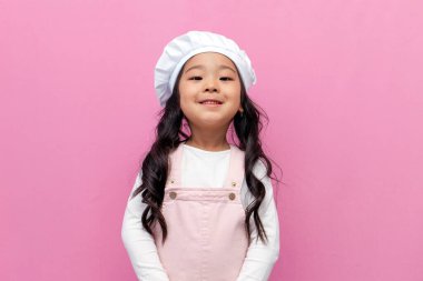 Aşçı üniformalı küçük Asyalı kız pembe izole arka planda gülümsüyor, Kore 'deki anaokulu çocuk aşçısı önlük ve şapka takıyor.