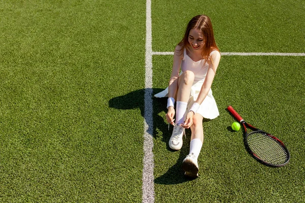 白いスポーツの若い女の子のテニス選手は彼女の靴を結ぶ上のスニーカーで緑のコート 女性コーチとテニスラケット コピースペース — ストック写真